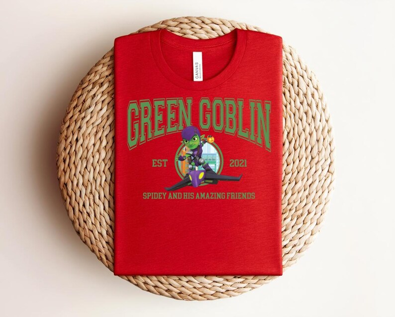 May include: A red t-shirt featuring the text "GREEN GOBLIN" in green arched lettering. Below the text is a cartoon image of the Green Goblin character and the text "SPIDEY AND HIS AMAZING FRIENDS". The shirt is folded on a woven surface.