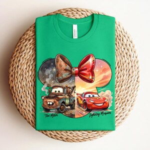 May include: A bright green t-shirt featuring a graphic of a bow shape containing two characters from the movie Cars. On the left, a brown rusty tow truck labelled Tow Mater. On the right, a red race car labelled Lightning McQueen.