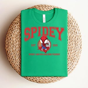 May include: Green t-shirt with the word "SPIDEY" in red arched lettering. Below the word "SPIDEY" is a cartoon image of Spider-Man in a circle, with the text "EST 2021" and "SPIDEY AND HIS AMAZING FRIENDS". The shirt is folded on a woven surface.