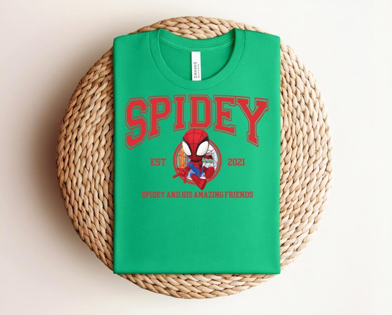 May include: Green t-shirt with the word "SPIDEY" in red arched lettering. Below the word "SPIDEY" is a cartoon image of Spider-Man in a circle, with the text "EST 2021" and "SPIDEY AND HIS AMAZING FRIENDS". The shirt is folded on a woven surface.