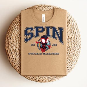 K&ouml;nnte beinhalten: Ein beiges T-Shirt mit dem Wort "SPIN" in marineblauen Buchstaben. Darunter befindet sich ein Cartoon-Superheldenbild. Der Text "EST 2021" und "SPIDEY AND HIS AMAZING FRIENDS" sind ebenfalls auf dem T-Shirt aufgedruckt.
