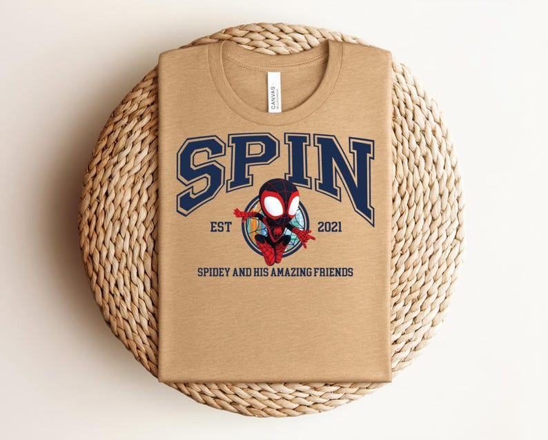 K&ouml;nnte beinhalten: Ein beiges T-Shirt mit dem Wort "SPIN" in marineblauen Buchstaben. Darunter befindet sich ein Cartoon-Superheldenbild. Der Text "EST 2021" und "SPIDEY AND HIS AMAZING FRIENDS" sind ebenfalls auf dem T-Shirt aufgedruckt.