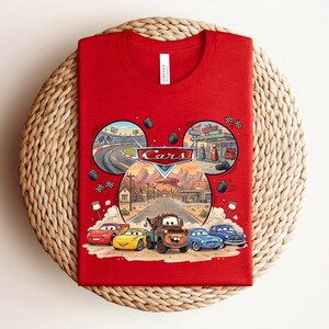 Puede incluir: Una camiseta roja con un estampado gr&aacute;fico del logotipo de Disney Pixar Cars y personajes como Rayo McQueen y Mate. El dise&ntilde;o est&aacute; dispuesto en forma de orejas de Mickey Mouse con escenas de un circuito de carreras y Radiator Springs.