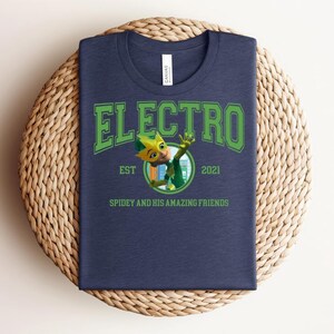 May include: Navy blue t-shirt with the word "ELECTRO" in green block letters. Below the word "ELECTRO" is a cartoon character in a circle, with the text "EST 2021" and "SPIDEY AND HIS AMAZING FRIENDS". The t-shirt is on a woven, circular surface.