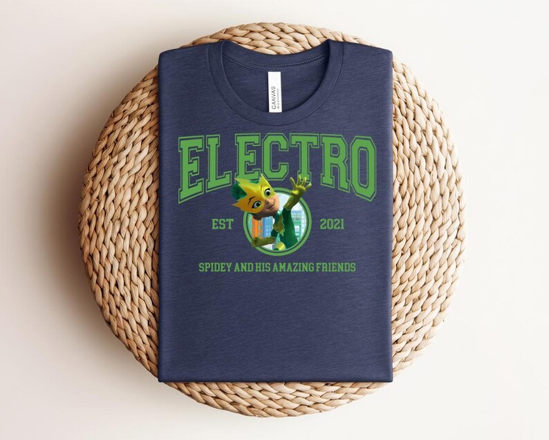 May include: Navy blue t-shirt with the word "ELECTRO" in green block letters. Below the word "ELECTRO" is a cartoon character in a circle, with the text "EST 2021" and "SPIDEY AND HIS AMAZING FRIENDS". The t-shirt is on a woven, circular surface.