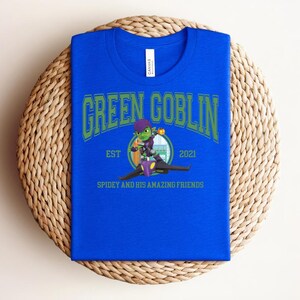 May include: A royal blue t-shirt with the words "GREEN GOBLIN" in green arched lettering. Below the text is a cartoon image of the Green Goblin character and the text "EST 2021" and "SPIDEY AND HIS AMAZING FRIENDS". The shirt is folded on a woven mat.