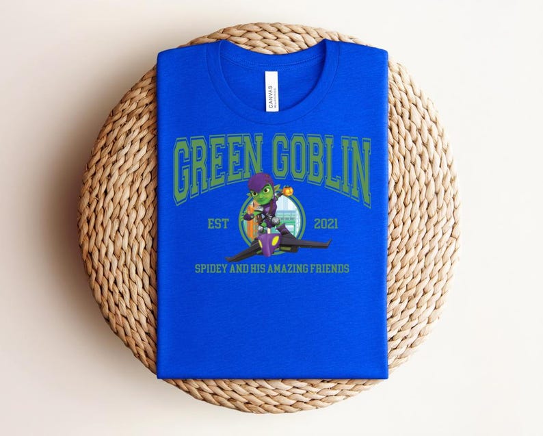 May include: A royal blue t-shirt with the words "GREEN GOBLIN" in green arched lettering. Below the text is a cartoon image of the Green Goblin character and the text "EST 2021" and "SPIDEY AND HIS AMAZING FRIENDS". The shirt is folded on a woven mat.