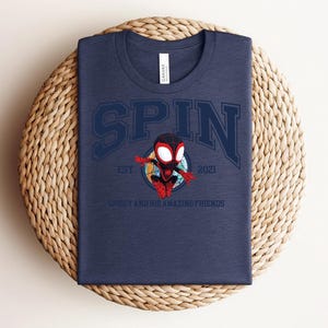 K&ouml;nnte beinhalten: Marineblaues T-Shirt mit dem Wort "SPIN" in einer College-Schriftart, mit einer Cartoon-Spidey-Figur. Das Shirt enth&auml;lt auch den Text "EST 2021" und "SPIDEY AND HIS AMAZING FRIENDS". Das Shirt ist auf einer gewebten, kreisf&ouml;rmigen Oberfl&auml;che gefaltet.