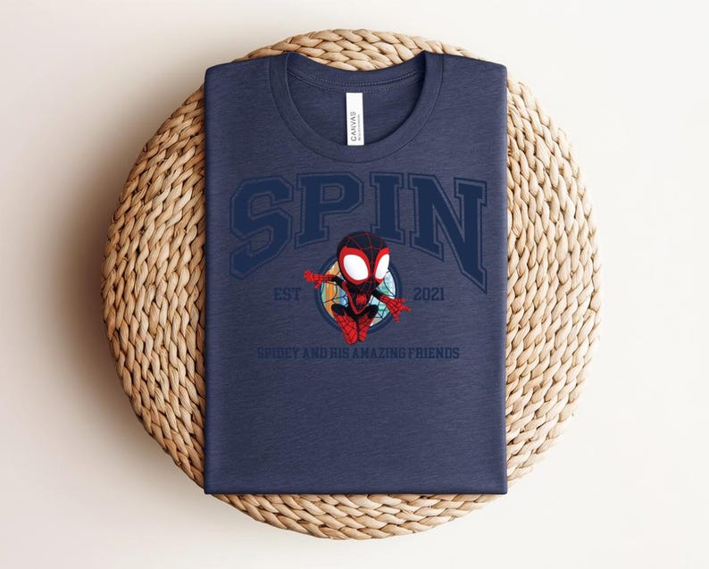 K&ouml;nnte beinhalten: Marineblaues T-Shirt mit dem Wort "SPIN" in einer College-Schriftart, mit einer Cartoon-Spidey-Figur. Das Shirt enth&auml;lt auch den Text "EST 2021" und "SPIDEY AND HIS AMAZING FRIENDS". Das Shirt ist auf einer gewebten, kreisf&ouml;rmigen Oberfl&auml;che gefaltet.