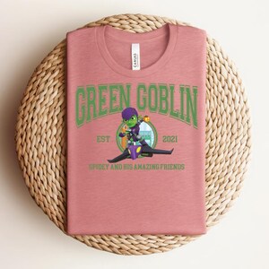 May include: A dusty rose t-shirt with a graphic of the Green Goblin riding a vehicle. The text "GREEN GOBLIN" arches across the top in green, with "EST 2021" below. The phrase "SPIDEY AND HIS AMAZING FRIENDS" is at the bottom.