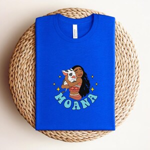 Puede incluir: Una camiseta azul real con una ilustraci&oacute;n gr&aacute;fica de un personaje de pelo largo y oscuro abrazando a un peque&ntilde;o cerdo manchado. El dise&ntilde;o incluye estrellas amarillas de cuatro puntas y el texto MOANA en letras azul claro.