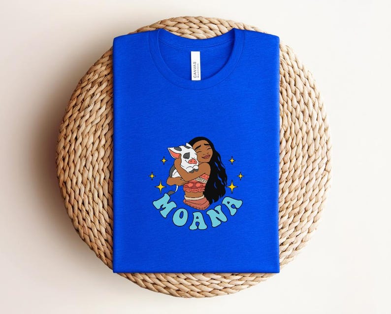 Puede incluir: Una camiseta azul real con una ilustraci&oacute;n gr&aacute;fica de un personaje de pelo largo y oscuro abrazando a un peque&ntilde;o cerdo manchado. El dise&ntilde;o incluye estrellas amarillas de cuatro puntas y el texto MOANA en letras azul claro.