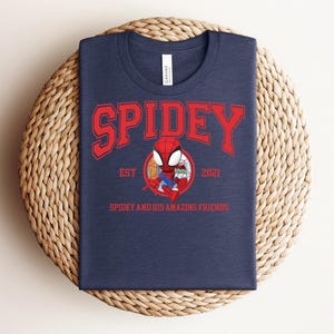 May include: Navy blue t-shirt with the word "SPIDEY" in red block letters. Below the word "SPIDEY" is a cartoon image of Spider-Man and the text "EST 2021" and "SPIDEY AND HIS AMAZING FRIENDS". The t-shirt is displayed on a woven, circular surface.