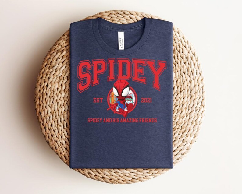 May include: Navy blue t-shirt with the word "SPIDEY" in red block letters. Below the word "SPIDEY" is a cartoon image of Spider-Man and the text "EST 2021" and "SPIDEY AND HIS AMAZING FRIENDS". The t-shirt is displayed on a woven, circular surface.