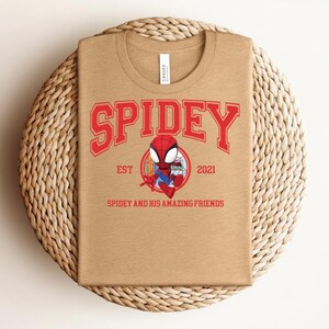 May include: A tan t-shirt featuring the word "SPIDEY" in red block letters above a cartoon image of Spider-Man. Below the image are the words "SPIDEY AND HIS AMAZING FRIENDS" and the year "2021". The shirt is displayed on a woven, circular placemat.