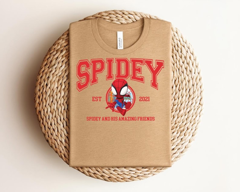 May include: A tan t-shirt featuring the word "SPIDEY" in red block letters above a cartoon image of Spider-Man. Below the image are the words "SPIDEY AND HIS AMAZING FRIENDS" and the year "2021". The shirt is displayed on a woven, circular placemat.