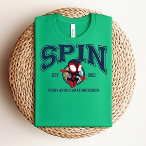 K&ouml;nnte beinhalten: Ein gr&uuml;nes T-Shirt mit dem Wort "SPIN" in marineblauen Blockbuchstaben mit wei&szlig;er Umrandung. Unter dem Wort "SPIN" befindet sich ein Cartoon-Bild eines Superhelden. Der Text "EST 2021" und "SPIDEY AND HIS AMAZING FRIENDS" sind ebenfalls auf dem T-Shirt aufgedruckt.