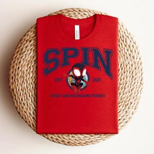 K&ouml;nnte beinhalten: Ein rotes T-Shirt mit dem Wort "SPIN" in marineblauen Blockbuchstaben. Unter dem Wort "SPIN" befindet sich ein Cartoon-Bild eines Superhelden. Der Text "EST 2021" und "SPIDEY AND HIS AMAZING FRIENDS" sind ebenfalls auf dem T-Shirt aufgedruckt.