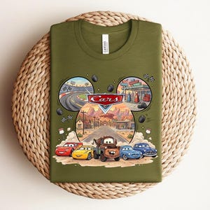 Puede incluir: Una camiseta verde oliva con un estampado gr&aacute;fico del logotipo de Disney Pixar Cars y personajes como Rayo McQueen, Mate y otros. El dise&ntilde;o incluye escenas circulares de una pista de carreras y una gasolinera sobre una carretera del desierto.