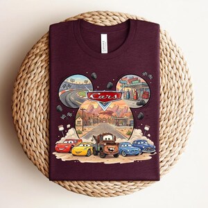 Puede incluir: Una camiseta de cuello redondo color granate con un gr&aacute;fico del logotipo y los personajes de Disney Pixar Cars, incluyendo a Rayo McQueen y Mate. El dise&ntilde;o tiene forma de cabeza de Mickey Mouse con escenas de carreras y Radiator Springs.