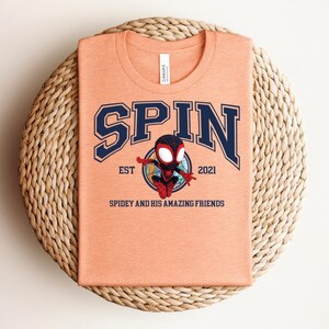 K&ouml;nnte beinhalten: Pfirsichfarbenes T-Shirt mit dem Wort "SPIN" in marineblauen College-Buchstaben. Darunter ist eine Cartoon-Figur von Spidey abgebildet, mit dem Text "EST 2021" und "Spidey and His Amazing Friends". Das T-Shirt ist auf einer gewebten Matte gefaltet.