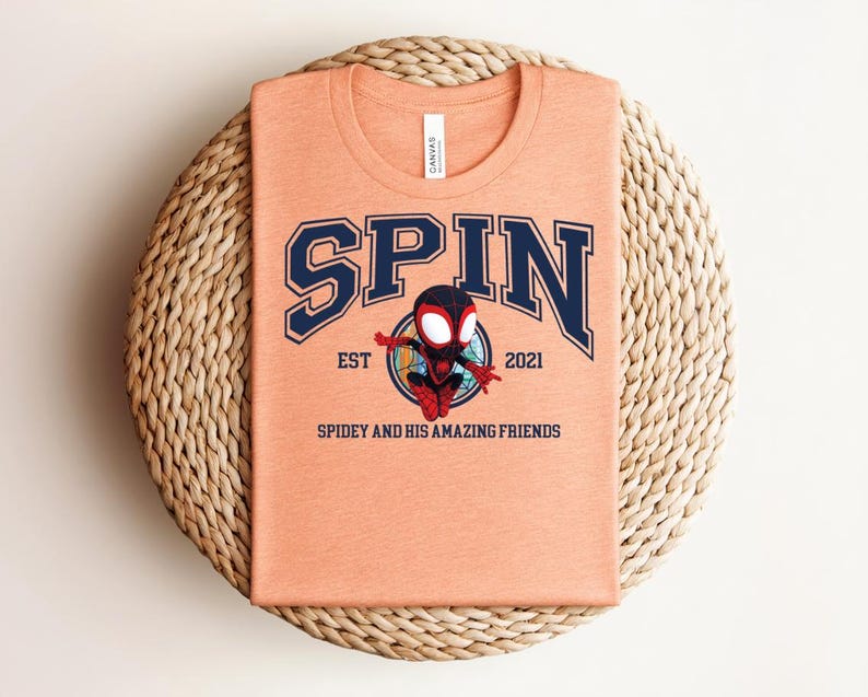 K&ouml;nnte beinhalten: Pfirsichfarbenes T-Shirt mit dem Wort "SPIN" in marineblauen College-Buchstaben. Darunter ist eine Cartoon-Figur von Spidey abgebildet, mit dem Text "EST 2021" und "Spidey and His Amazing Friends". Das T-Shirt ist auf einer gewebten Matte gefaltet.