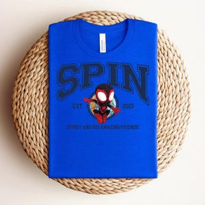 K&ouml;nnte beinhalten: K&ouml;nigsblaues T-Shirt mit dem Wort "SPIN" in marineblauen Blockbuchstaben. Darunter befindet sich ein Cartoon-Bild eines Superhelden und der Text "EST 2021" und "SPIDEY AND HIS AMAZING FRIENDS". Das Shirt liegt auf einer gewebten Matte.