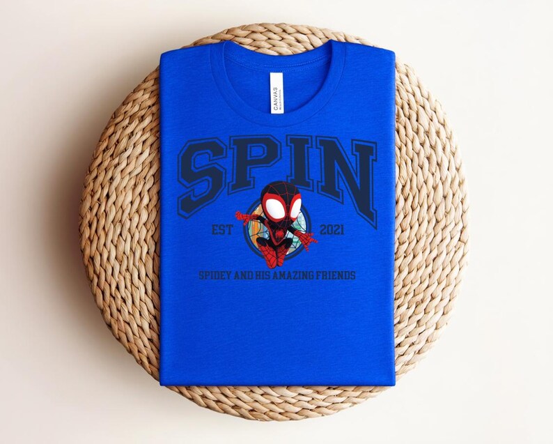 K&ouml;nnte beinhalten: K&ouml;nigsblaues T-Shirt mit dem Wort "SPIN" in marineblauen Blockbuchstaben. Darunter befindet sich ein Cartoon-Bild eines Superhelden und der Text "EST 2021" und "SPIDEY AND HIS AMAZING FRIENDS". Das Shirt liegt auf einer gewebten Matte.