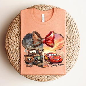 May include: A peach-coloured t-shirt featuring a graphic of a bow shape containing Tow Mater on the left and Lightning McQueen on the right. The text Tow Mater and Lightning McQueen is printed below the respective characters in a black script font.