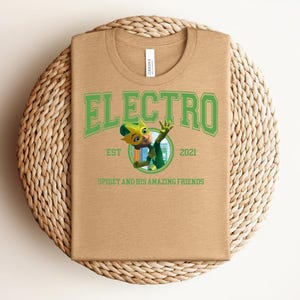 May include: A tan t-shirt with a green graphic that says "ELECTRO" above a cartoon character and the text "SPIDEY AND HIS AMAZING FRIENDS". The shirt has a crew neck and is folded on a woven mat.