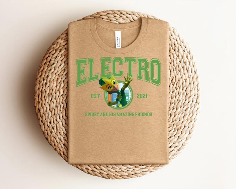 May include: A tan t-shirt with a green graphic that says "ELECTRO" above a cartoon character and the text "SPIDEY AND HIS AMAZING FRIENDS". The shirt has a crew neck and is folded on a woven mat.