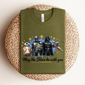Pode incluir: Uma t-shirt de gola redonda verde-azeitona com uma impress&atilde;o gr&aacute;fica de BB-8, o Mandaloriano, Grogu e Darth Vader ao lado de um ca&ccedil;a estelar X-wing. O texto "May the Force be with you" est&aacute; impresso em letra cursiva preta sob as figuras.