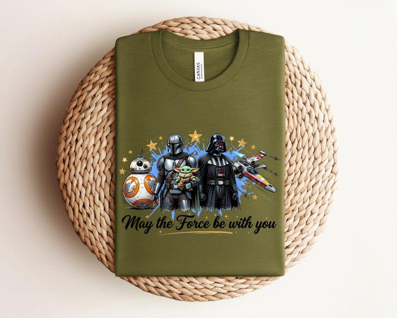 Pode incluir: Uma t-shirt de gola redonda verde-azeitona com uma impress&atilde;o gr&aacute;fica de BB-8, o Mandaloriano, Grogu e Darth Vader ao lado de um ca&ccedil;a estelar X-wing. O texto "May the Force be with you" est&aacute; impresso em letra cursiva preta sob as figuras.