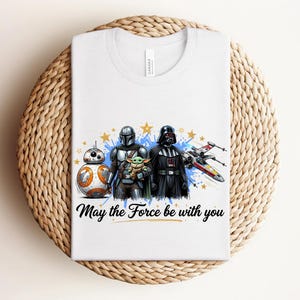 Pode incluir: Uma t-shirt branca com uma impress&atilde;o gr&aacute;fica de BB-8, o Mandaloriano, Grogu e Darth Vader ao lado de um ca&ccedil;a estelar X-wing. O design inclui salpicos de tinta azul, estrelas amarelas e o texto "May the Force be with you" em preto.