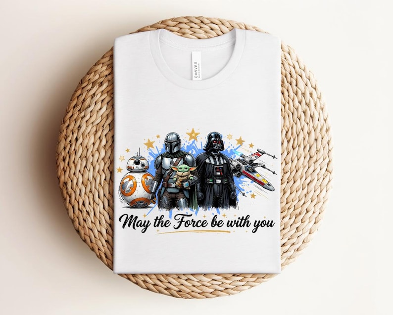 Pode incluir: Uma t-shirt branca com uma impress&atilde;o gr&aacute;fica de BB-8, o Mandaloriano, Grogu e Darth Vader ao lado de um ca&ccedil;a estelar X-wing. O design inclui salpicos de tinta azul, estrelas amarelas e o texto "May the Force be with you" em preto.