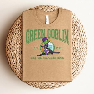 May include: A tan t-shirt featuring the text "GREEN GOBLIN" in green, with a cartoon image of the Green Goblin riding a glider. The text "EST 2021" and "SPIDEY AND HIS AMAZING FRIENDS" are also printed on the shirt.