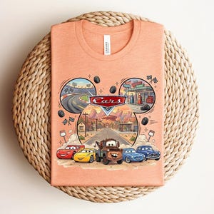Puede incluir: Una camiseta color melocot&oacute;n con un gr&aacute;fico del logotipo de Disney Pixar Cars y personajes como Rayo McQueen, Mate y otros. El dise&ntilde;o est&aacute; dispuesto en forma de orejas de Mickey Mouse con escenas de una pista de carreras y Radiator Springs.