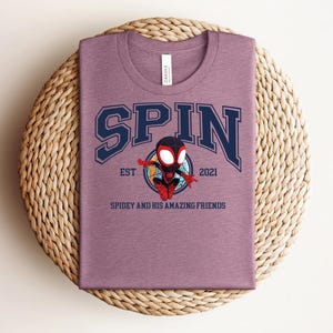 K&ouml;nnte beinhalten: Ein mauvefarbenes T-Shirt mit dem Wort "SPIN" in marineblauen Buchstaben. Darunter befindet sich ein Cartoon-Bild von Spidey, mit dem Text "EST 2021" und "SPIDEY AND HIS AMAZING FRIENDS".