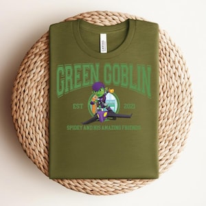 May include: Olive green t-shirt featuring the text "GREEN GOBLIN" in green arched lettering above an illustration of the Green Goblin character riding a glider. The text "EST 2021" and "SPIDEY AND HIS AMAZING FRIENDS" are also visible.