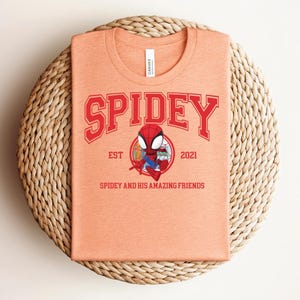 May include: A peach-colored t-shirt featuring the word "SPIDEY" in red block letters above a cartoon image of Spider-Man. The shirt also includes the text "EST 2021" and "SPIDEY AND HIS AMAZING FRIENDS". The shirt is folded on a woven, circular placemat.