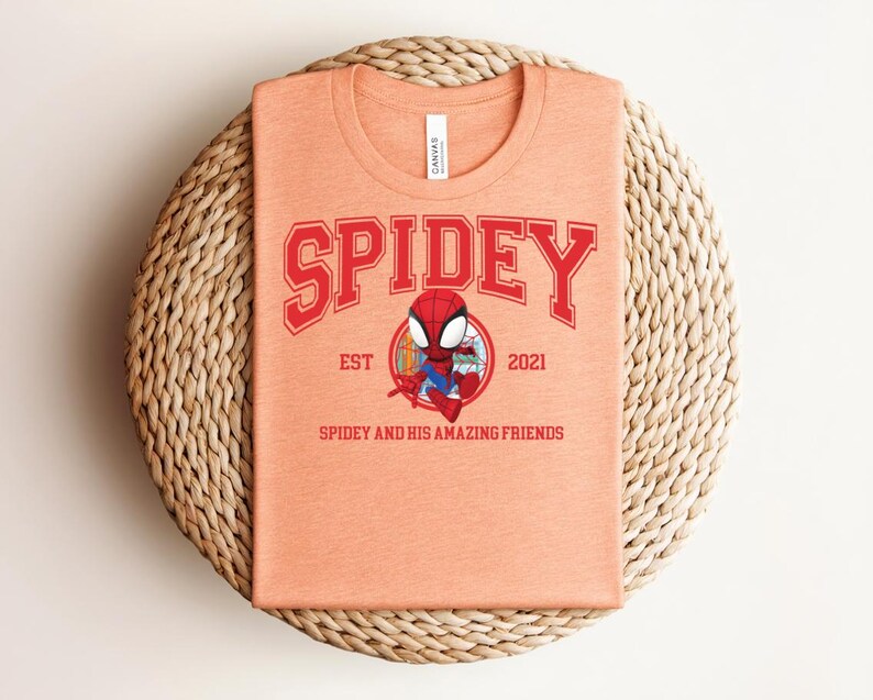May include: A peach-colored t-shirt featuring the word "SPIDEY" in red block letters above a cartoon image of Spider-Man. The shirt also includes the text "EST 2021" and "SPIDEY AND HIS AMAZING FRIENDS". The shirt is folded on a woven, circular placemat.