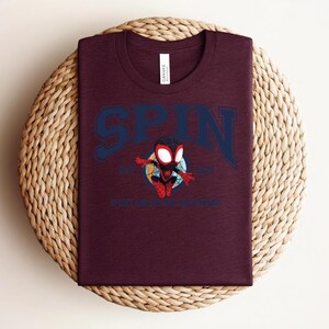 K&ouml;nnte beinhalten: Burgunderfarbenes T-Shirt mit dem Wort "SPIN" in Marineblau, das sich oben bogenf&ouml;rmig erstreckt. Darunter befindet sich eine Cartoon-Grafik eines Superhelden. Der Text "SPIDER-VERSE" steht darunter. Das T-Shirt wird auf einer gewebten, runden Matte pr&auml;sentiert.