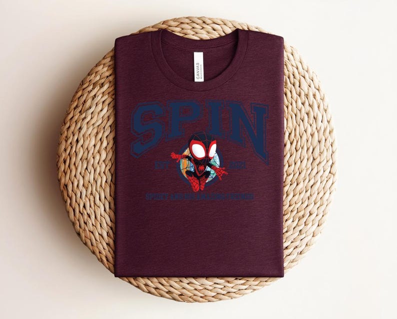 K&ouml;nnte beinhalten: Burgunderfarbenes T-Shirt mit dem Wort "SPIN" in Marineblau, das sich oben bogenf&ouml;rmig erstreckt. Darunter befindet sich eine Cartoon-Grafik eines Superhelden. Der Text "SPIDER-VERSE" steht darunter. Das T-Shirt wird auf einer gewebten, runden Matte pr&auml;sentiert.
