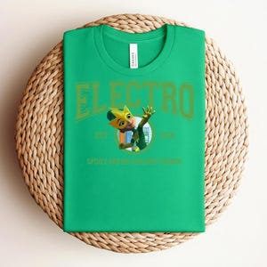 May include: A bright green t-shirt with the word "ELECTRO" in a green, collegiate-style font. Below the word is a cartoon character and the text "EST 1963 SPIDEY AND HIS AMAZING FRIENDS". The shirt is folded on a woven mat.