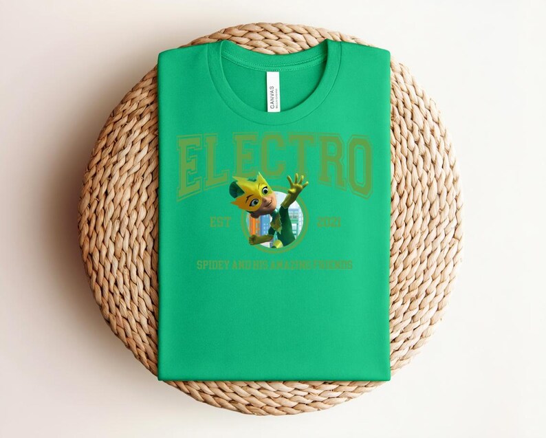 May include: A bright green t-shirt with the word "ELECTRO" in a green, collegiate-style font. Below the word is a cartoon character and the text "EST 1963 SPIDEY AND HIS AMAZING FRIENDS". The shirt is folded on a woven mat.