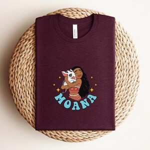 Puede incluir: Una camiseta de color granate oscuro con cuello redondo que presenta una ilustraci&oacute;n gr&aacute;fica de un personaje de pelo largo y oscuro abrazando a un peque&ntilde;o cerdo blanco y negro. La palabra MOANA est&aacute; impresa en letras azul claro debajo.