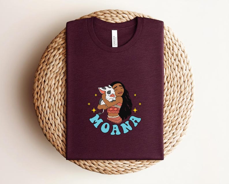 Puede incluir: Una camiseta de color granate oscuro con cuello redondo que presenta una ilustraci&oacute;n gr&aacute;fica de un personaje de pelo largo y oscuro abrazando a un peque&ntilde;o cerdo blanco y negro. La palabra MOANA est&aacute; impresa en letras azul claro debajo.