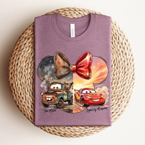 May include: A mauve crew neck t-shirt featuring a graphic of Tow Mater and Lightning McQueen beneath a large red and brown bow. The design includes the text Tow Mater and Lightning McQueen in a script font, set against a circular background.