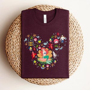 K&ouml;nnte beinhalten: Ein weinrotes T-Shirt mit einer farbenfrohen Grafik von Arielle, der Meerjungfrau, angeordnet in Form eines Mickey Mouse-Kopfes. Das Design umfasst verschiedene Charaktere und Elemente aus dem Film, wie Fische, Blumen und das Schloss.