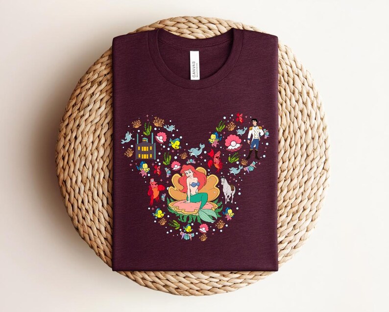 K&ouml;nnte beinhalten: Ein weinrotes T-Shirt mit einer farbenfrohen Grafik von Arielle, der Meerjungfrau, angeordnet in Form eines Mickey Mouse-Kopfes. Das Design umfasst verschiedene Charaktere und Elemente aus dem Film, wie Fische, Blumen und das Schloss.
